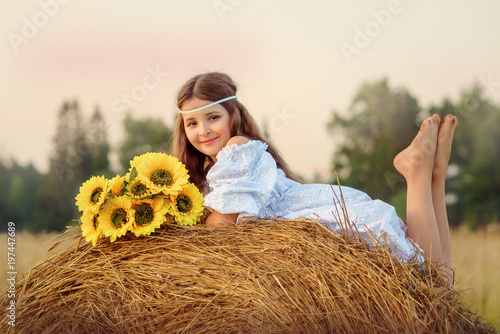 Девочка в белом платье летом в поле лежит на стоге сена с букетом подсолнухов