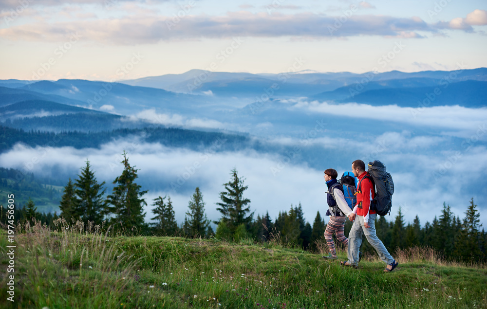 Landscape of mighty mountains with a slight haze and tourists with backpacks walking holding hands along a mountain path. The concept of a healthy active traveling lifestyle