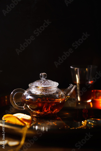 Small glass teapot and glasses with hot black tea, dried rose petals, pocket magnifier on golden chain, squeezed orange slice on golden tray. Evening light. Side view.