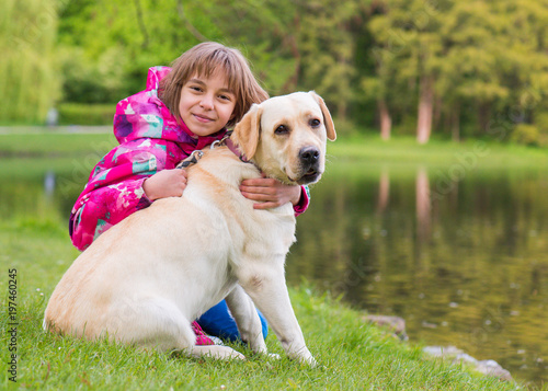 Little girl with labrador retriever on walk in park. Child sitting on green grass with dog - outdoor in nature portrait. Pet  domestic animal and people concept.