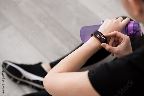 A woman's hand with a smart watch. Bottle with water in hand. Close-up. Sitting on the floor. Black sportswear. Looks time. Black sneakers with white soles. Wooden floor.