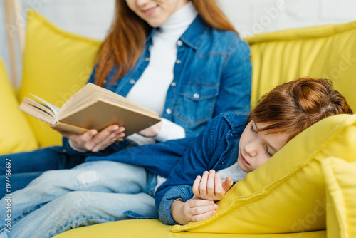 Mother reading book to sleeping daughter on yellow sofa