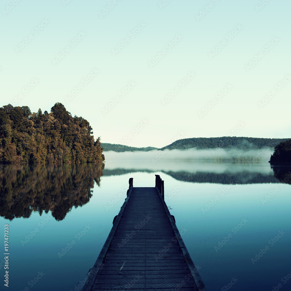 Naklejka premium Obraz minimalistycznego niebieskiego krajobrazu przystani obok pięknego spokojnego jeziora. W tej scenie jest kilka drzew liściastych i zamglonych gór. Krajobraz odbija się w wodzie.