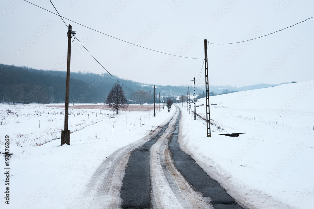 snowy winter landscape, asphalt road leads in to the hills, pylons and wires