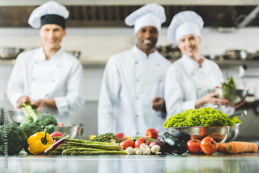 smiling multicultural chefs at restaurant kitchen with vegetables on foreground