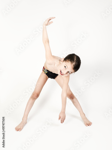 A cute boy with long hair gathered in a tail in swimming trunks, engaged in gymnastic exercises. Light background