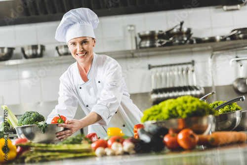 smiling attractive chef taking bowl with vegetables and looking at camera at restaurant kitchen photo