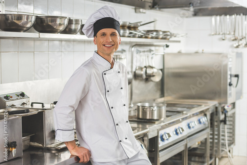 smiling chef leaning on table and looking at camera at restaurant kitchen