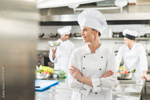 smiling chef standing with crossed arms and looking away at restaurant kitchen