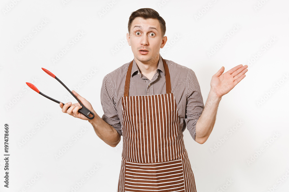 Young man chef in striped brown apron, shirt holding black kitchen serving plastic pair of tongs for salad isolated on white background. Male housekeeper or houseworker. Kitchenware, cuisine concept. Young man chef in striped brown apron, shirt holding black kitchen serving plastic pair of tongs for salad isolated on white background. Male housekeeper or houseworker. Kitchenware, cuisine concept.