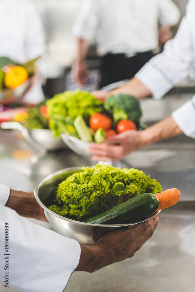 cropped image of multiethnic chefs holding bowls with vegetables at restaurant kitchen cropped image of multiethnic chefs holding bowls with vegetables at restaurant kitchen