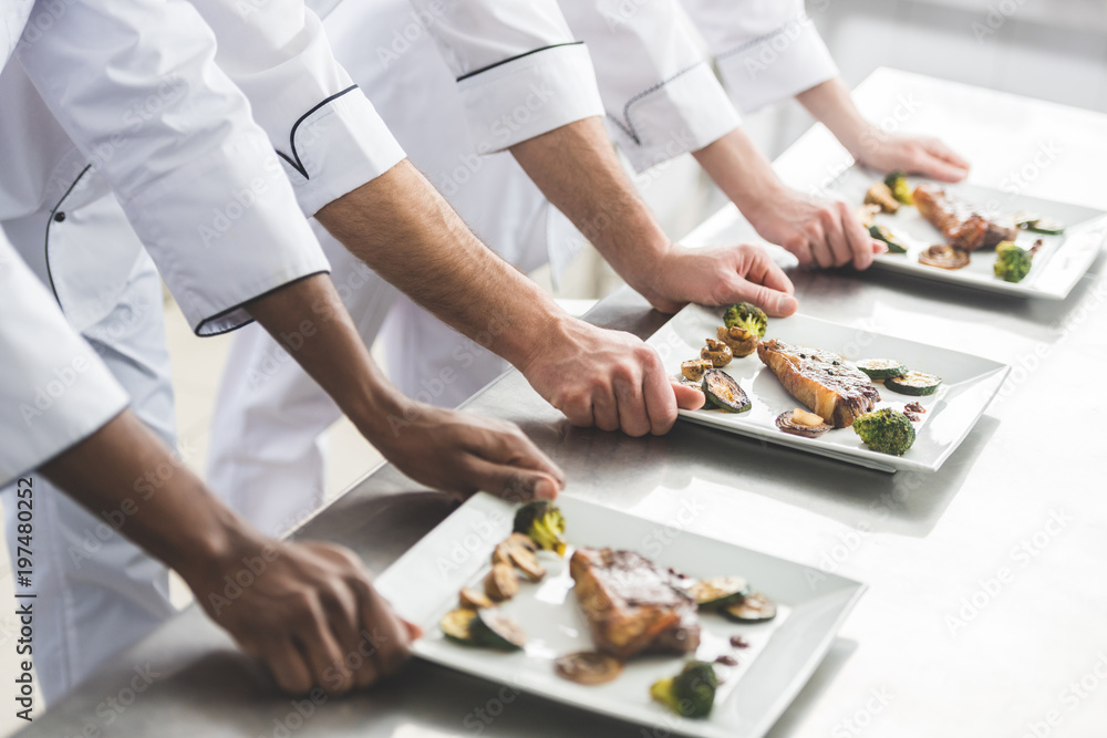 cropped image of multicultural chefs with plates with steaks at restaurant kitchen