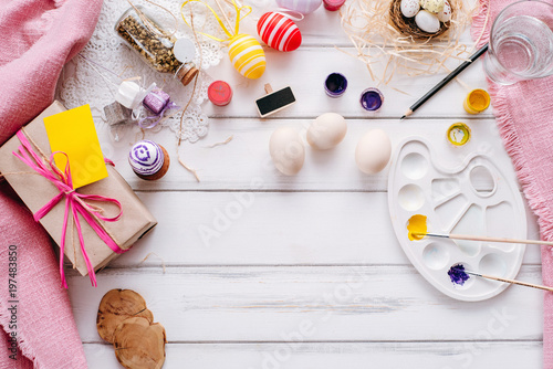Top view of handmade easter objects laying on white wooden desk, daughter's hands painting, decorate eggs, holding brushed. Day before Easter, child painting eggs for Easter