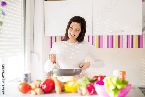 Young cheerful gorgeous woman is preparing breakfast while holding pan and standing in front of a kitchen counter filled with fresh vegetables.