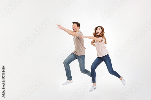 Full-length picture of delighted couple man and woman in casual t-shirt and jeans running and pointing fingers on copyspace with smile, isolated over white background