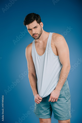 handsome bearded young man in shorts and white singlet looking at camera on blue