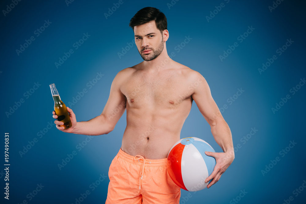 handsome young man in shorts holding bottle of summer drink and inflatable ball and looking at camera on blue
