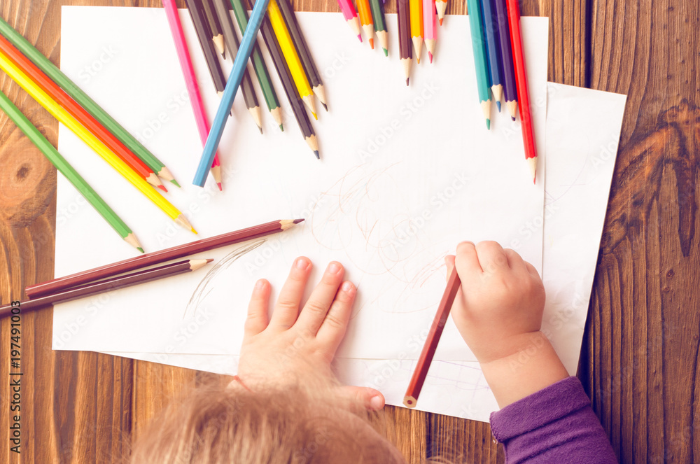 The child's hands are painted with colored pencils on a white sheet of paper on a wooden table The child's hands are painted with colored pencils on a white sheet of paper on a wooden table