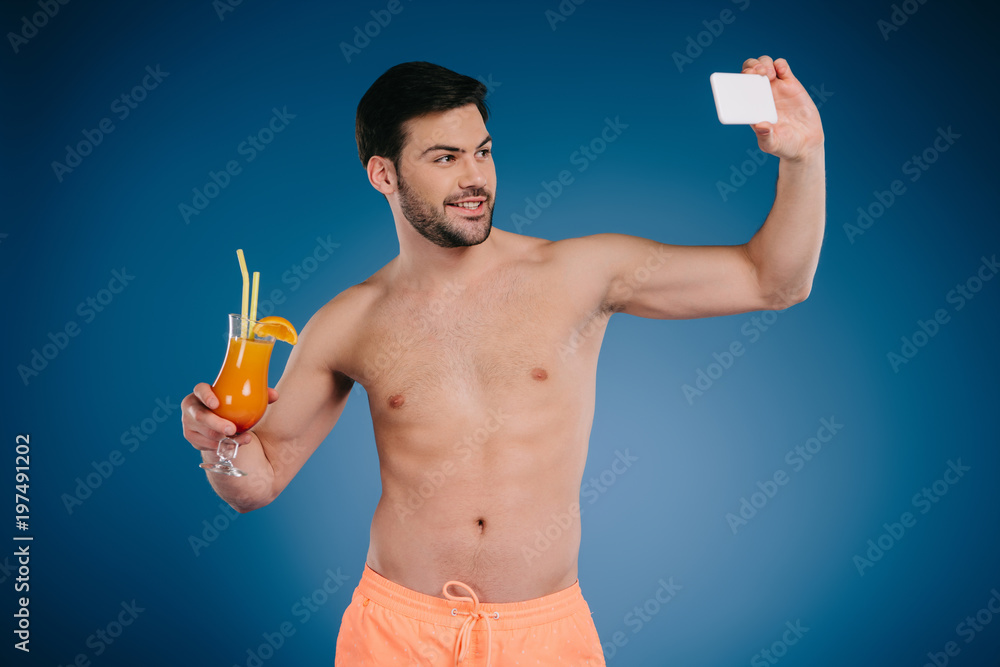 handsome shirtless young man in shorts holding glass of summer cocktail and taking selfie with smartphone on blue handsome shirtless young man in shorts holding glass of summer cocktail and taking selfie with smartphone on blue