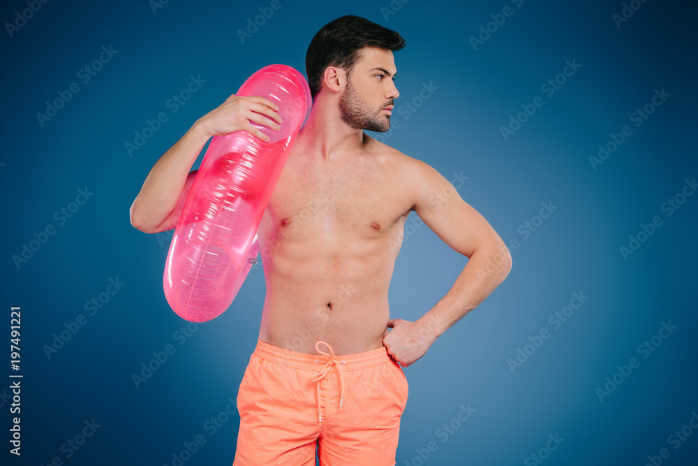 handsome young man in shorts holding inflatable ring and looking away on blue