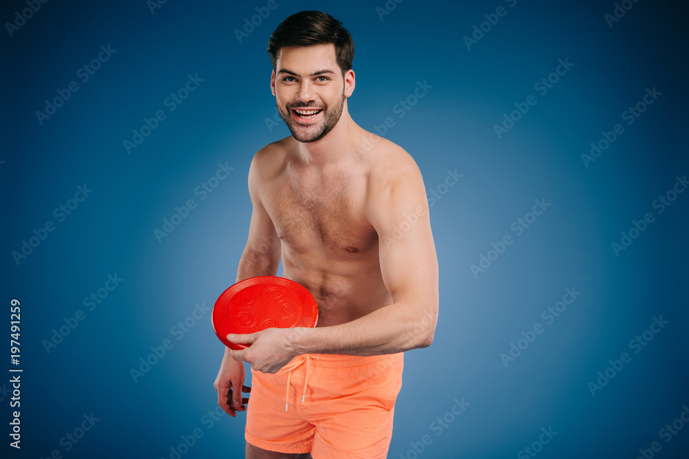 handsome young bare chested man in shorts holding flying disk and smiling at camera on blue