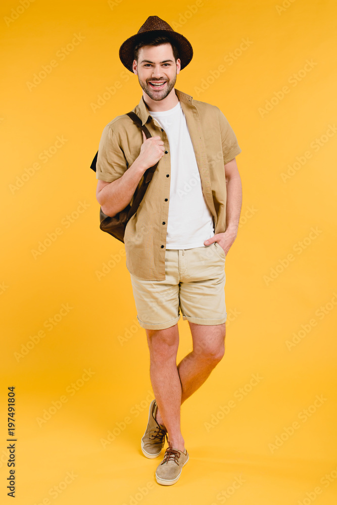 full length view of handsome young man in shorts and hat holding backpack and smiling at camera isolated on yellow full length view of handsome young man in shorts and hat holding backpack and smiling at camera isolated on yellow