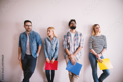 Group of young stylish people leaning against the wall bored before a job interview with folders in hands in the waiting room.