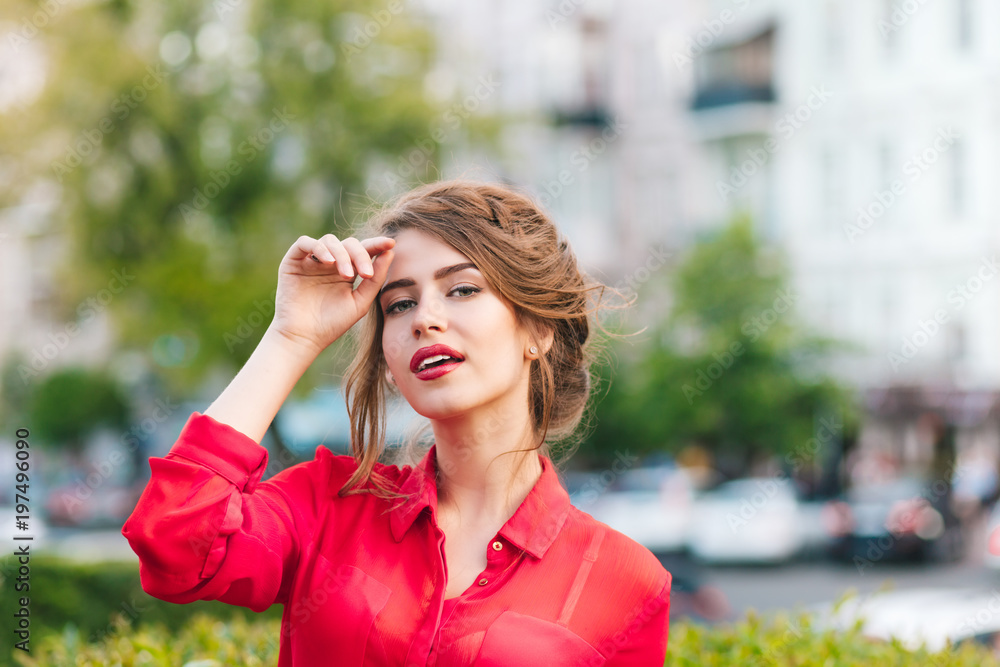 Close-up horizontal portrait of pretty girl standing in park. She wears red blouse and nice hairstyle. She is looking to the camera. Close-up horizontal portrait of pretty girl standing in park. She wears red blouse and nice hairstyle. She is looking to the camera.