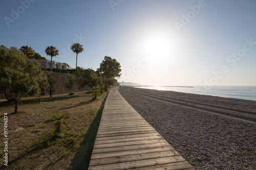 landscape at dawn of wooden walkway in Els Terrers Beach, in Benicassim, Castellon, Valencia, Spain, Europe. Palm trees, buildings, blue clear sky and Mediterranean Sea
 photo