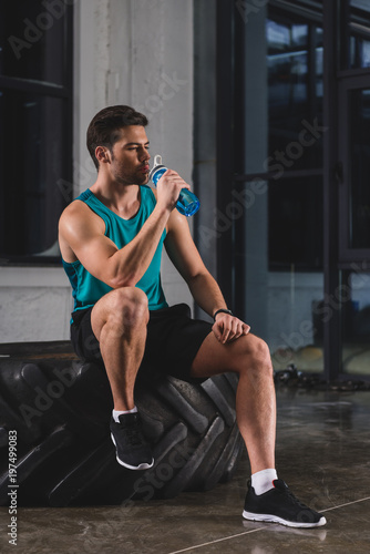 sportsman sitting on tire and drinking water after cross training in gym