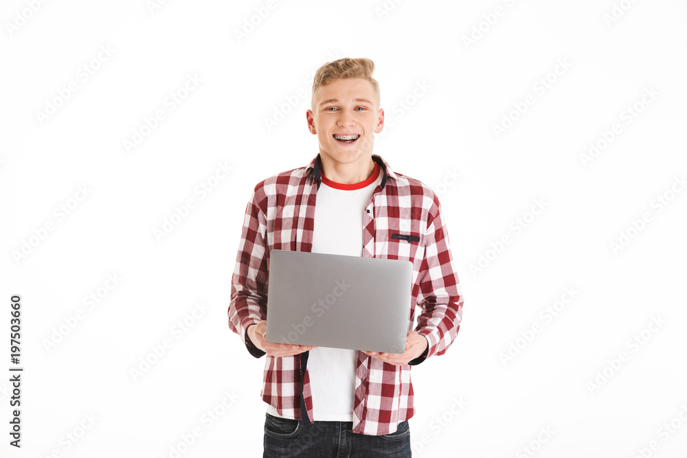 Portrait of smiling smart teenage boy wearing plaid shirt and braces studying using silver laptop, isolated over white background Portrait of smiling smart teenage boy wearing plaid shirt and braces studying using silver laptop, isolated over white background