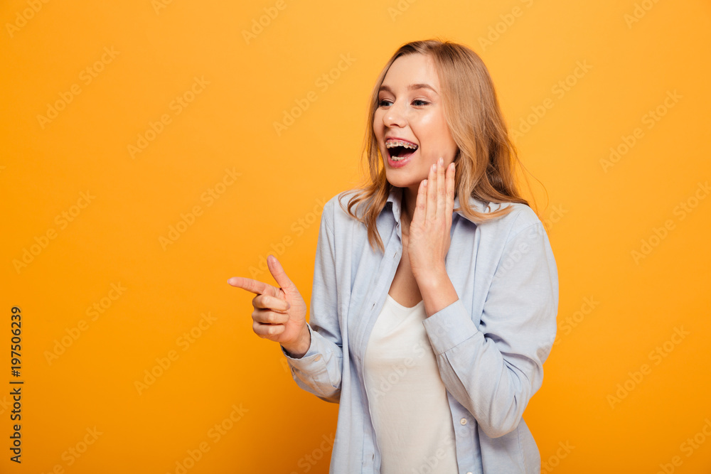 Portrait of delighted woman with auburn hair smiling and pointing index finger aside on copyspace text or product, isolated over yellow background Portrait of delighted woman with auburn hair smiling and pointing index finger aside on copyspace text or product, isolated over yellow background
