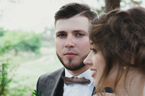 Beautiful wedding couple portrait in the forest. The bride with elegant hairstyle is standing near the bearded groom in bow tie. Hipster rustic stylish love story outdoors.