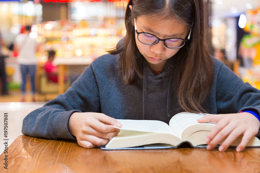 Girl with glasses reading a thick fiction story book at a table inside a public place with blurred shops background, Girl with glasses reading a thick fiction story book at a table inside a public place with blurred shops background,