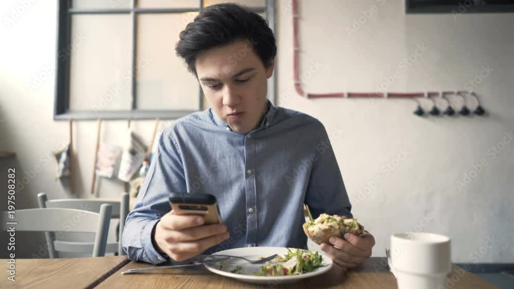 Young teenager with smartphone eating hamburger sitting in cafe
