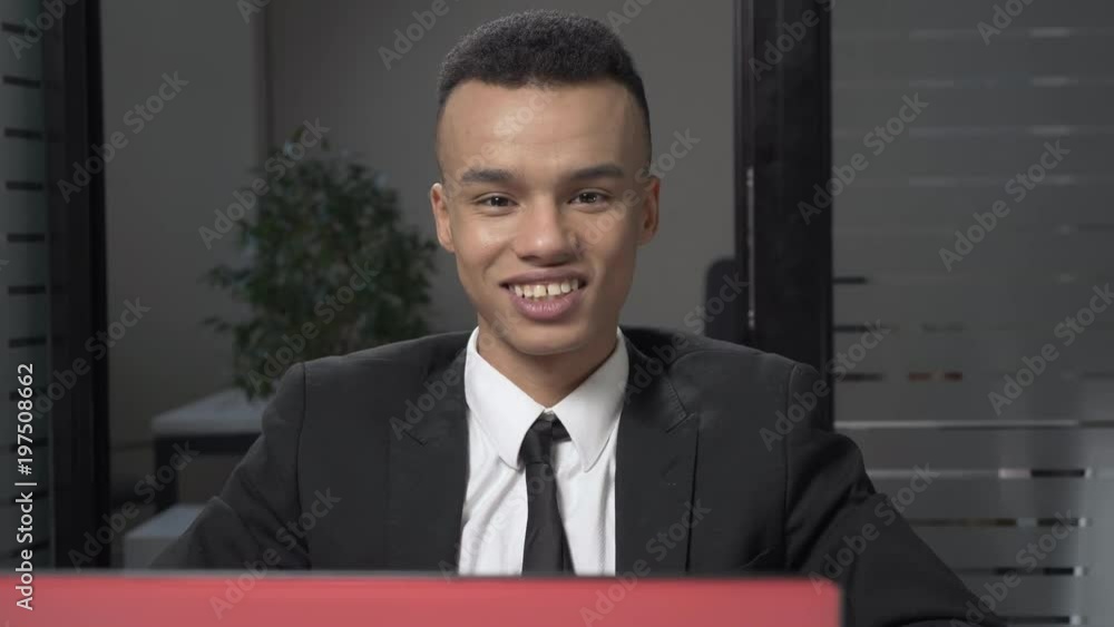 Young successful African businessman sitting in office, working on a laptop, looking at camera and smiling, portrait concept. 60 fps Young successful African businessman sitting in office, working on a laptop, looking at camera and smiling, portrait concept. 60 fps