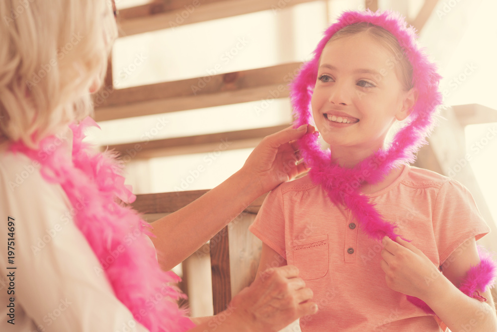 Having fun. Happy positive cute girl smiling and looking at her grandmother while wearing a pink feather boa