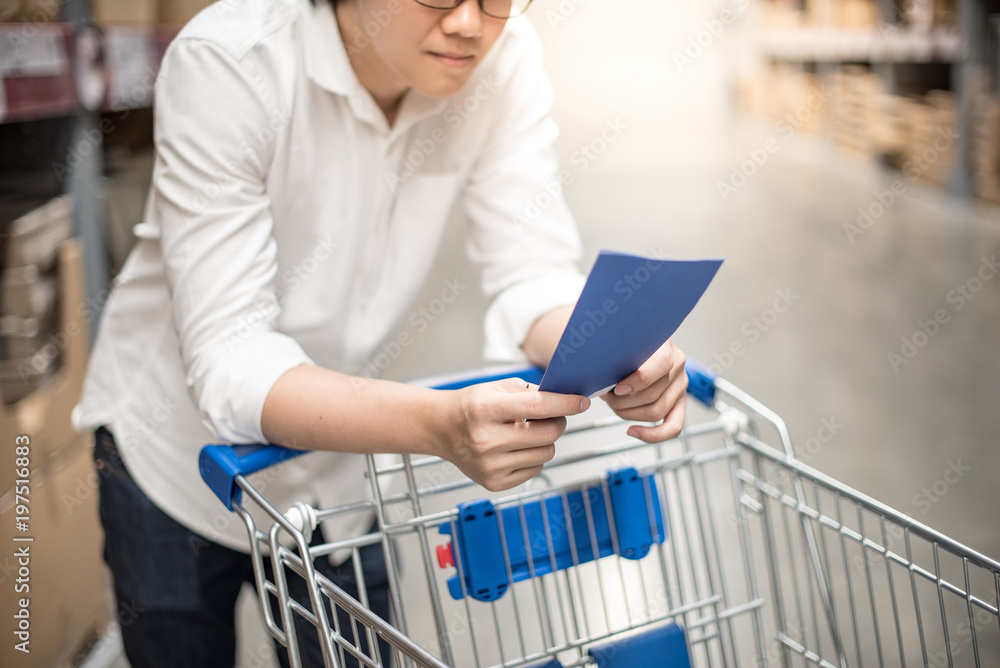Young Asian man standing with cart checking the shopping list in warehouse wholesale, shopping warehousing concept Young Asian man standing with cart checking the shopping list in warehouse wholesale, shopping warehousing concept