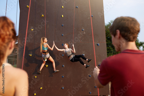 Smiling girls giving high five hanging on ropes at artificial climbing training wall and insured by friends on belaying harness. Rock climbing safety system. Smiling women practicing in bouldering.