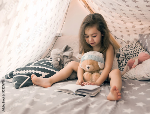 Cute happy little girl playing with toys and reading book in teepee and bed. Close up photo of happy child