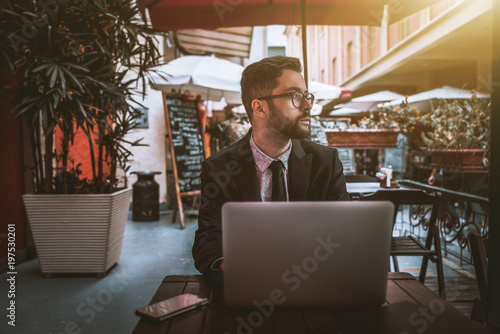 Young diffident bearded businessman in the glasses and a formal suit is sitting with the laptop in a street bar alone and thoughtfully looking aside; unconfident man entrepreneur with netbook in cafe