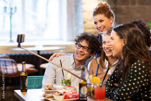 leisure  technology  friendship  people and holidays concept - happy friends with drinks and taking picture by smartphone selfie stick at bar or cafe