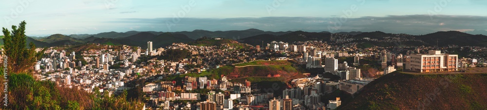 Wide panoramic view from a high point an urban landscape lit by bright evening sun: plenty of residential, office, educational houses, hills with favelas, mountain ridge in the distance; Juiz de Fora