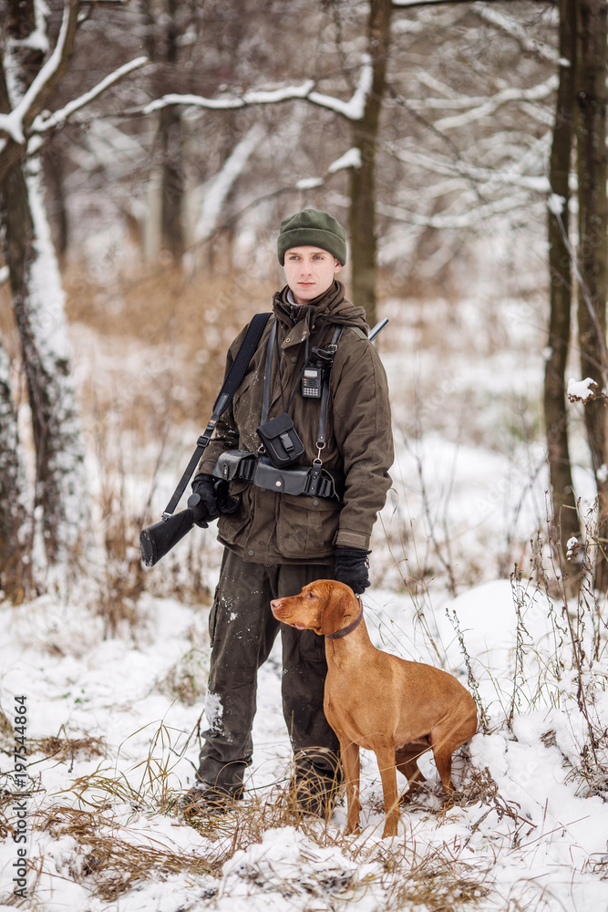 Male hunter in camouflage, armed with a rifle, standing in a snowy winter forest with duck prey