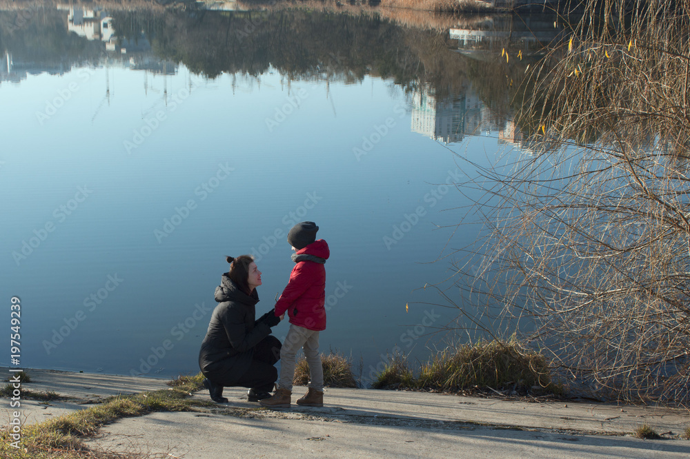 On the shore of lake mom talks to her son. Family walking in the park near lake. Mother and son look at each other in spring. buildings are reflected in water