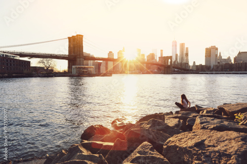 Blurred of a girl sitting on rocks with the view of Brooklyn Bridge and Manhattan skyline at sunset in vintage colour