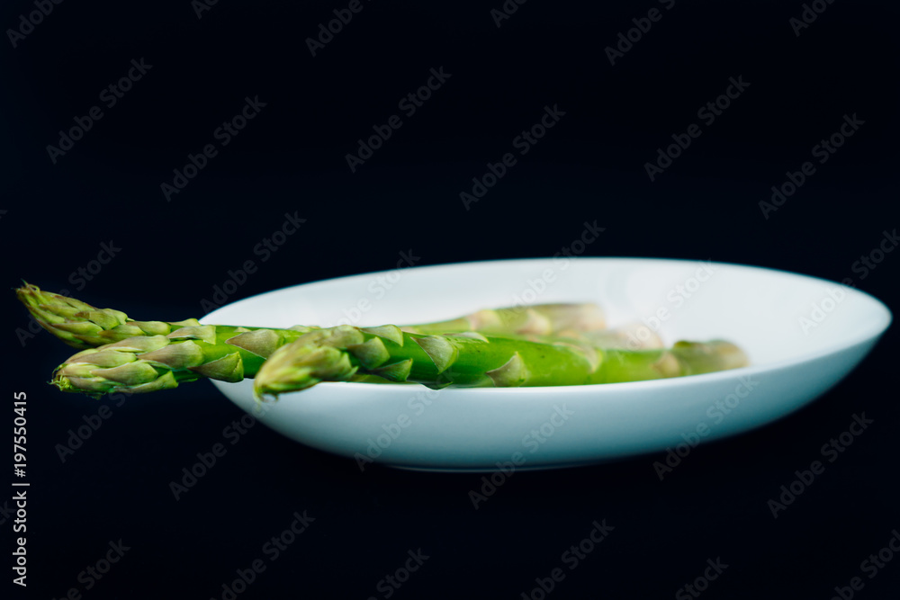 Asparagus, or garden asparagus, sparrow-grass in white plate on black background Asparagus, or garden asparagus, sparrow-grass in white plate on black background