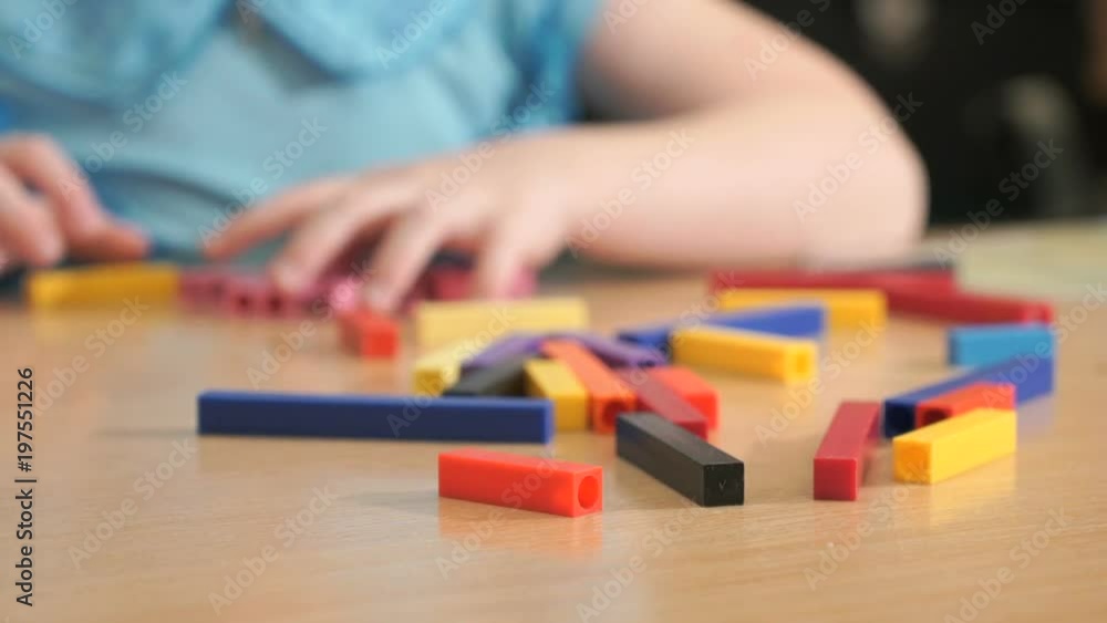 Close-up of hands of unidentified little girl in blue shirt sitting at desk in the kindergarten playing in developing designer - making figures of stripes of various colors. Close-up of hands of unidentified little girl in blue shirt sitting at desk in the kindergarten playing in developing designer - making figures of stripes of various colors.