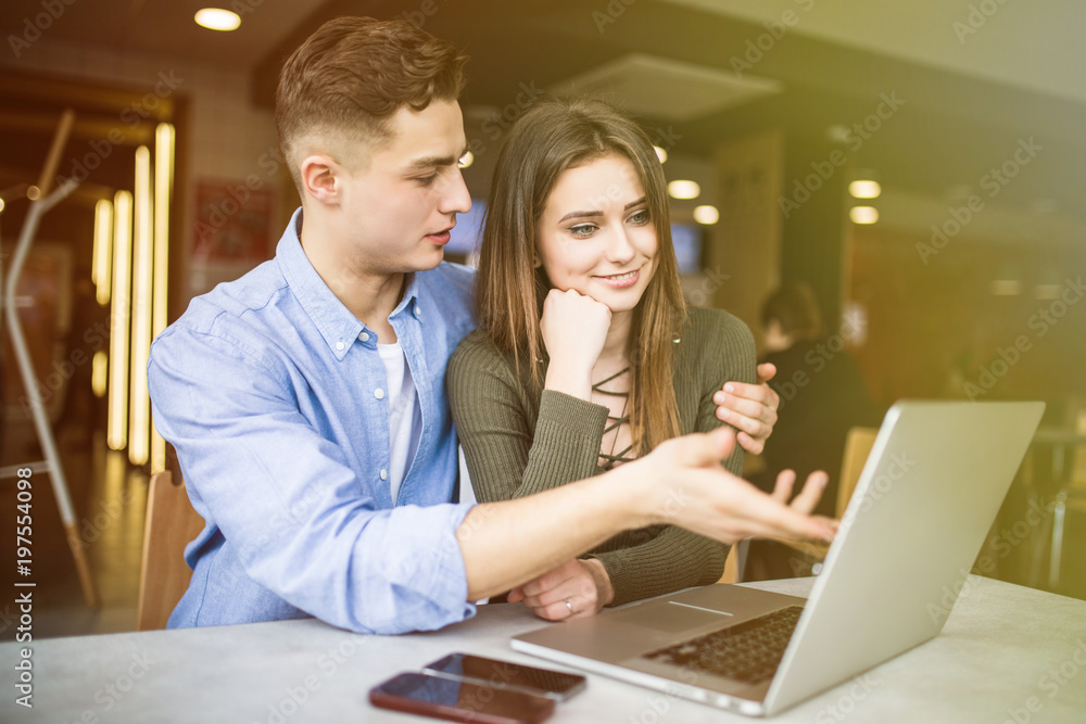 Fototapeta premium Happy young couple is using a laptop, drinking coffee and smiling while sitting at the cafe