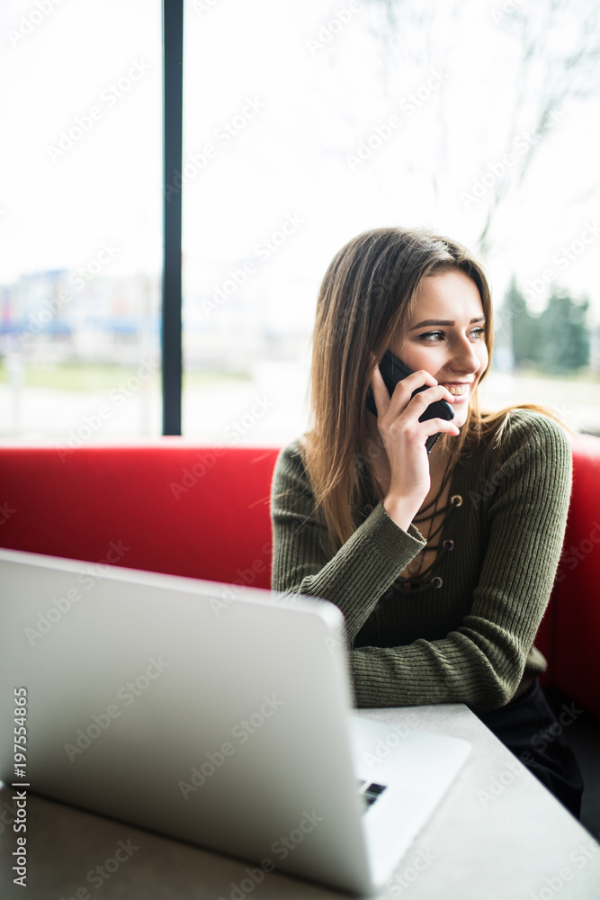 Happy woman using laptop and phone in cafe. Young beautiful girl sitting in a coffee shop and working on computer Happy woman using laptop and phone in cafe. Young beautiful girl sitting in a coffee shop and working on computer